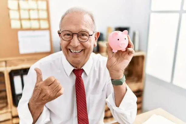 Photo of Senior business man holding piggy bank pointing thumb up to the side smiling happy with open mouth