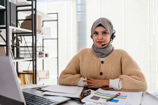 Muslim business woman with headset working in office. Happy arab woman working in company service center wearing headphone and using computer helping solving client problem. Islamic business woman working on computer at call center with copy space.