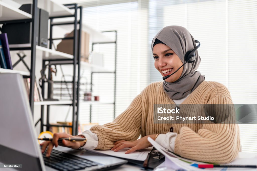 Black Islamic Lady In Hijab And Headset Having Video Call On Laptop Muslim business woman with headset working in office. Happy arab woman working in company service center wearing headphone and using computer helping solving client problem. Islamic business woman working on computer at call center with copy space. Customer Service Representative Stock Photo