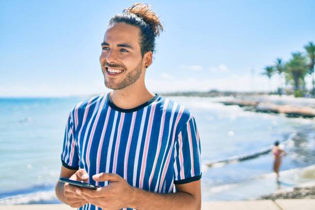 Young hispanic man smiling happy using smartphone at the beach.