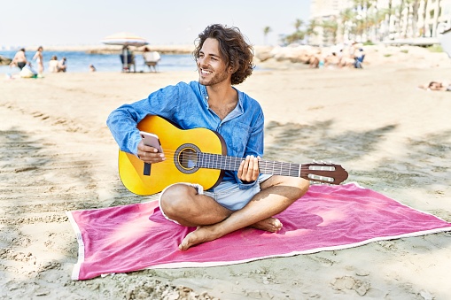 Young hispanic man playing classical guitar sitting on sand at the beach.