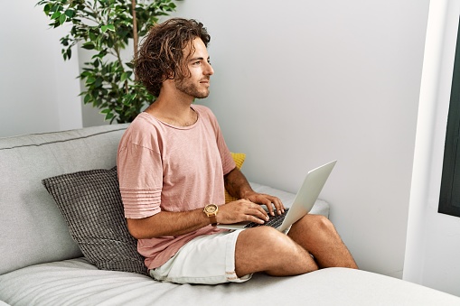 Young hispanic man sitting on the sofa at home using laptop looking to side, relax profile pose with natural face and confident smile.