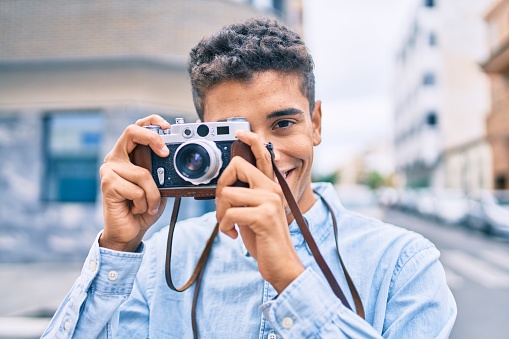 Young latin tourist man smiling happy using vintage camera walking at the city.