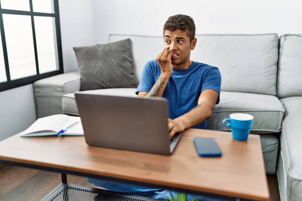 Young handsome hispanic man using laptop sitting on the floor looking stressed and nervous with hands on mouth biting nails. anxiety problem. Young handsome hispanic man using laptop sitting on the floor looking stressed and nervous with hands on mouth biting nails. anxiety problem. nail biting stock pictures, royalty-free photos & images