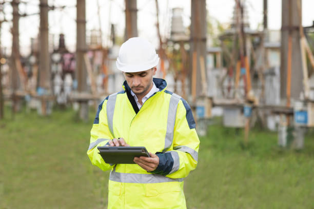 istock Man standing near high voltage substation and typing on device. Male engineer in uniform and helmet standing outdoor and tapping on tablet 1346579926
