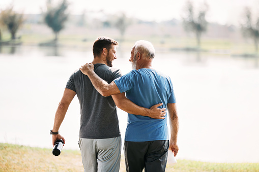 Rear view of athletic father and son talking while walking embraced by the lake.