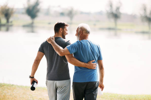 Rear view of athletic father and son talking while walking embraced by the lake. Back view of happy athletic man and his senior father walking embraced while exercising in nature. Copy space. adult stock pictures, royalty-free photos & images