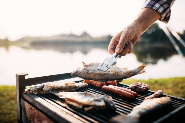 primer plano del hombre asando pescado en la parrilla de barbacoa mientras acampa junto al río. - prepared fish fish grilled close up fotografías e imágenes de stock