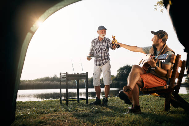 Happy senor man and his son toasting with beer while camping by the lake. Happy man playing acoustic guitar and toasting with his father who is grilling meat during their camping day. father and son guitar stock pictures, royalty-free photos & images