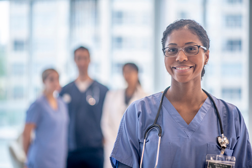 A female nurse of African decent stands in the hallway posing for a portrait.  She is wearing a blue scrubs and has a stethoscope on as her colleagues stand in the background.