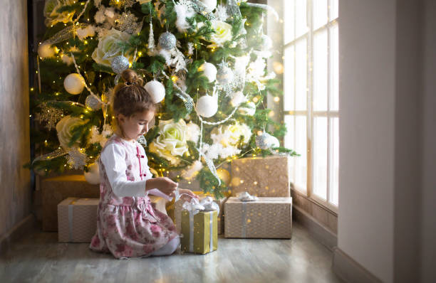 маленькая девочка в красивом платье сидит под елкой с подарочной коробкой и бантом. свет из большого окна, темная комната, домашний уют, рож� - christmas window magic house стоковые фото и изображения