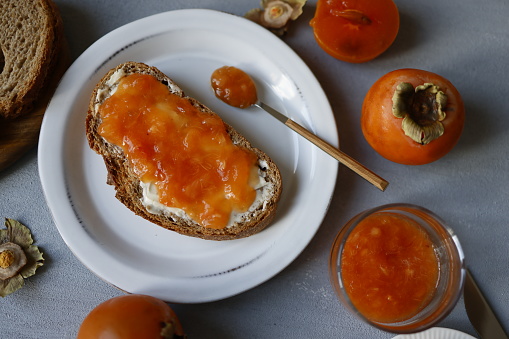 Istanbul, Turkey-September 30, 2021: Persimmon paste spread on greasy bread on white plate on gray rough concrete floor. There is persimmon paste in a dessert spoon on the plate. There is also a bowl of persimmon paste, shells and whole persimmons in a bowl around the plate.