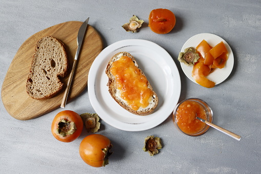 Istanbul, Turkey-September 30, 2021: Persimmon paste spread on greasy bread on white plate on gray rough concrete floor. There is persimmon paste in a dessert spoon on the plate. There is also a bowl of persimmon paste, shells and whole persimmons in a bowl around the plate.