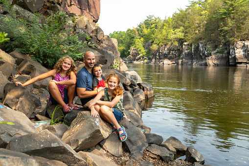 A father with his three children smiling for the camera. They are sitting on rocks on the bank of the St. Croix River between Minnesota & Wisconsin, USA.