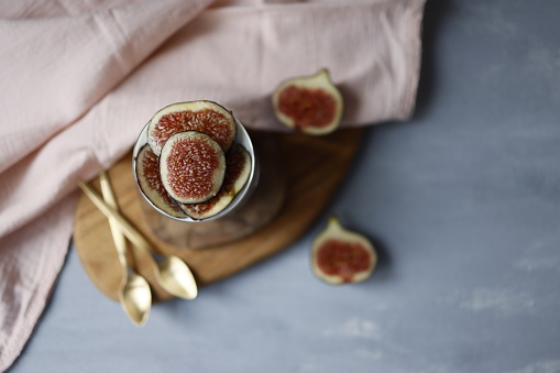 Istanbul, Turkey-September 29, 2021: Oatmeal yogurt garnished with fresh fig slices in a round glass cup on a wooden serving on a gray rough concrete floor. There is a light pink linen cloth napkin with lace around the glass cup and two gold colored dessert spoons. Yogurt and oatmeal lined up in layers in a cup. There are slices of fresh figs cut in half on the floor. Shot with Canon EOS R5.