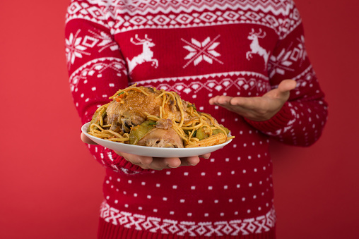 Cropped closeup photo of young man in red and white christmas sweater demonstrating large plate of food on isolated red background