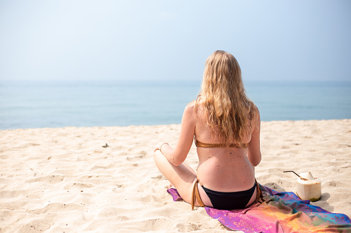 A woman with long blond hair sits in a lotus position on the sandy seashore in Thailand. Travel and tourism.