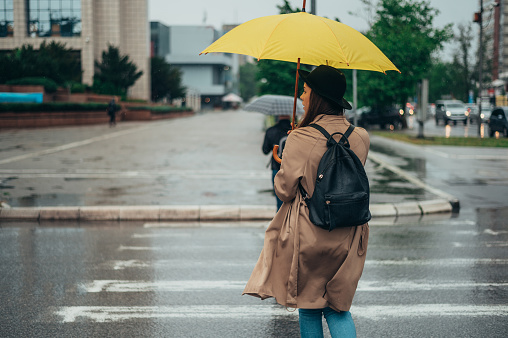 Young beautiful woman walking on the crossroad during rain and holding a yellow umbrella dressed in a coat