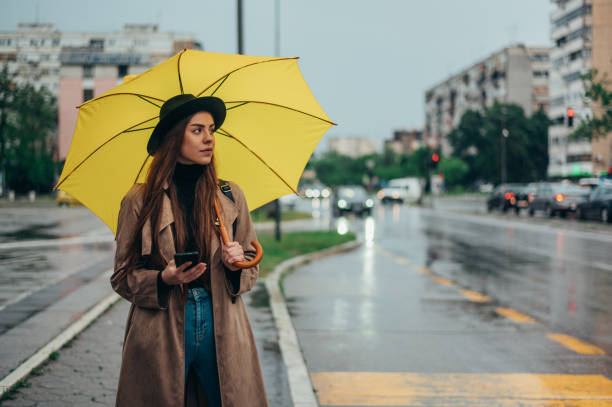 スマートフォンを使って黄色い傘を持つ若い美しい女性 - umbrella women storm yellow ストックフォトと画像