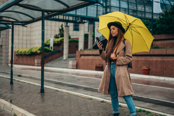 istock Woman holding a yellow umbrella and standing on a bus stop while using a smartphone 1346618775