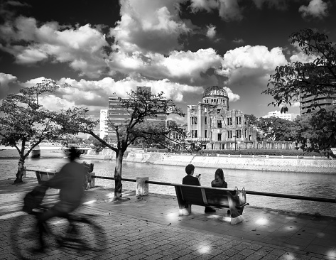 Hiroshima, Hiroshima Prefecture, Japan - October 9, 2021: A cyclist passes a couple sitting next to the Motoyasu River by the Genbaku Dome, or Atomic Bomb Dome, near the ground zero hypocenter.