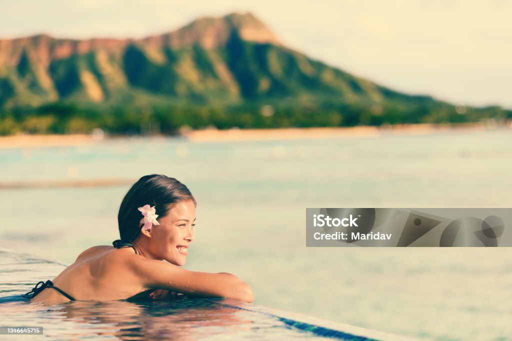 Smiling Woman Relaxing In Infinity Swimming Pool Smiling beautiful woman relaxing in infinity swimming pool. Side view of happy female looking at ocean view on sunny day. Young tourist is enjoying summer vacation against mountain. Hawaii Islands Stock Photo