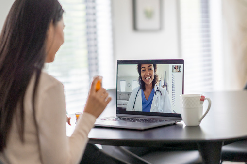 A female patient of Asian decent sits at her kitchen table with a laptop out as she has an appointment with her doctor remotely.  She is dressed casually and holding out her medication as she asks her doctor about it.  The doctor can be seen on the screen in a white lab coat as she talks with the patient.