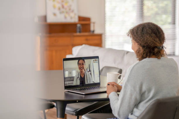 Patient Meeting Remotely with her Doctor A female patent of African decent meets with her doctor remotely via a video call.  She is sitting at her kitchen table with her laptop out and a cup of coffee on the table with her.  The doctor can be seen on the screen wearing a white lab coat as she talks with her patient. telemedicine stock pictures, royalty-free photos & images