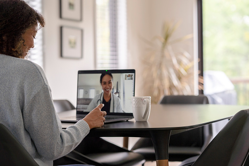 A female patent of African decent meets with her doctor remotely via a video call.  She is sitting at her kitchen table with her laptop out and her prescription on the table with her.  The doctor can be seen on the screen wearing a white lab coat as she talks with her patient.