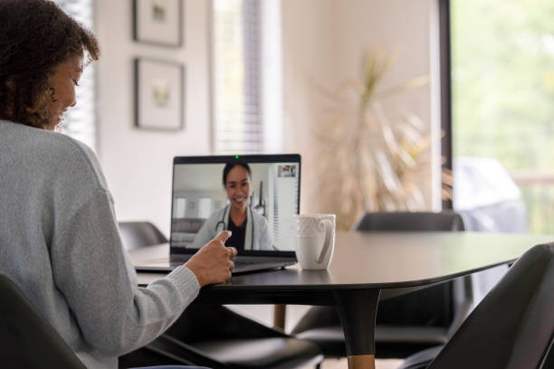 Patient Meeting Remotely with her Doctor A female patent of African decent meets with her doctor remotely via a video call.  She is sitting at her kitchen table with her laptop out and her prescription on the table with her.  The doctor can be seen on the screen wearing a white lab coat as she talks with her patient. virtual event stock pictures, royalty-free photos & images