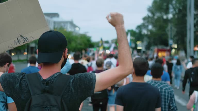 Waving arm and fist, protest activist marches in riot crowd with banner in hand.