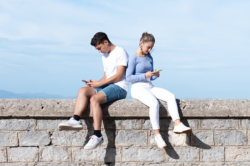 Sad teenager couple concentrated on their phones sitting on a stone wall. Digital addiction and dopamine concept.