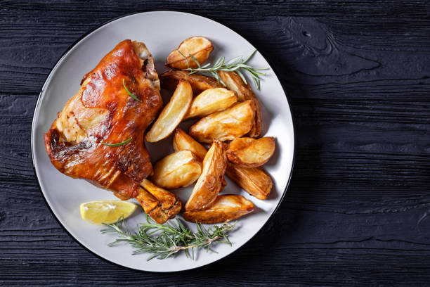 knusprig gebratene schweineschäfte auf einem teller mit kartoffelspalten, rosmarin, zitrone auf einem schwarzen holztisch, horizontale ansicht von oben, flache lage, freiraum - pork hock stock-fotos und bilder