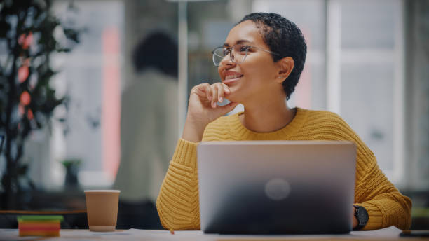 feliz especialista en marketing latino en gafas trabajando en computadora portátil en oficina creativa ocupada. hermosa y diversa gerente de proyecto dudando y pensando antes de responder un correo electrónico a un colega. - hesitating fotografías e imágenes de stock