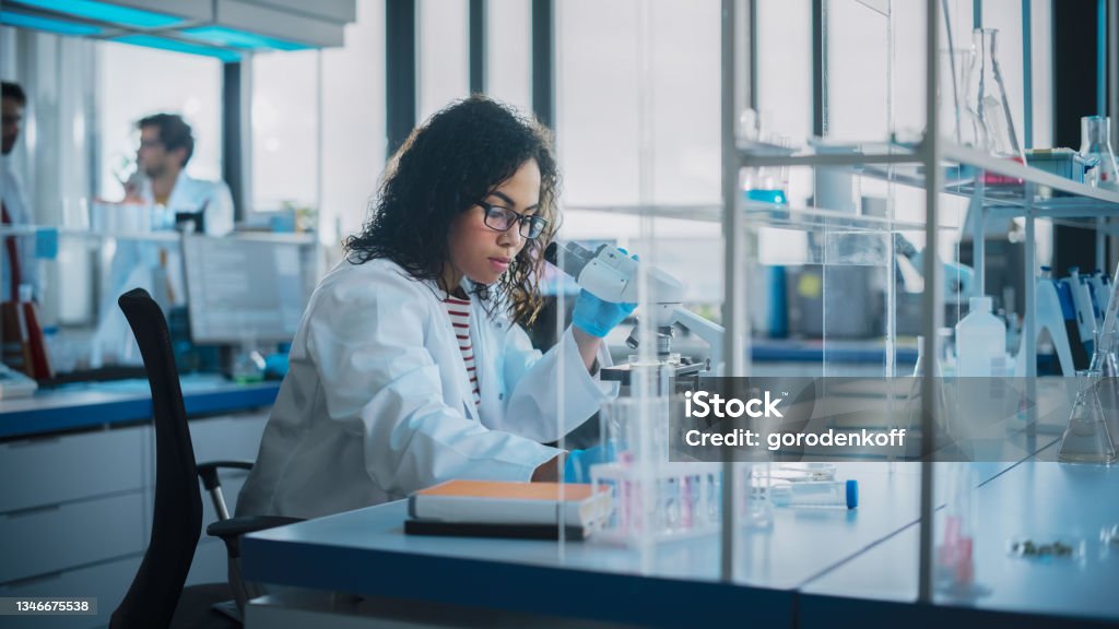 Medical Science Laboratory: Portrait of Beautiful Black Scientist is Using Microscope Does Analysis of Test Sample. Ambitious Young Biotechnology Specialist, working with Advanced Equipment Science Stock Photo