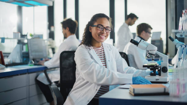 medical science laboratory: beautiful black scientist benutzt mikroskop, schaut in die kamera und lächelt charmant. junger biotechnologie-wissenschaftsspezialist mit technologisch fortschrittlichen geräten. - charmingly stock-fotos und bilder