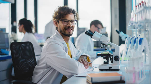 medical science laboratory: der hübsche latinische wissenschaftler benutzt das mikroskop, schaut in die kamera und lächelt charmant. junger biotechnologiespezialist mit technisch fortschrittlichen geräten. - charmingly stock-fotos und bilder