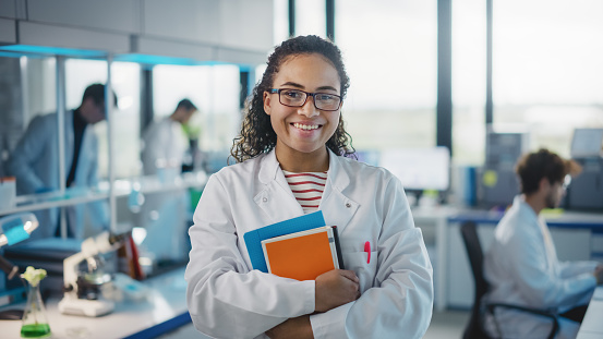 Laboratorio de Ciencias Médicas: Hermosa joven científica negra inteligente con bata blanca y gafas, sostiene libros de prueba, sonríe mirando a la cámara. Equipo diverso de especialistas. Retrato medio tomado photo