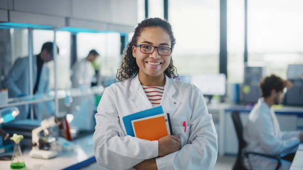 medical science laboratory: beautiful smart young black scientist wearing white coat and glasses, holds test books, smiles looking at camera. diverse team of specialists. medium portrait shot - biologisch fotos stockfoto's en -beelden