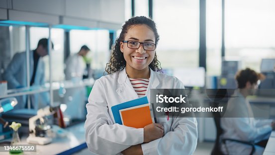 istock Medical Science Laboratory: Beautiful Smart Young Black Scientist Wearing White Coat and Glasses, Holds Test Books, Smiles Looking at Camera. Diverse Team of Specialists. Medium Portrait Shot 1346675550