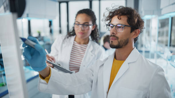 istock Medical Science Laboratory: Handsome Latin Male Scientist Writes Detailed Project Data Analysis on the Board, His Black Female Colleague Talks. Young Scientists Solving Problems. 1346675587