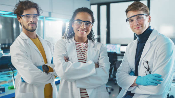 medical science laboratory with team of three young successful scientists. beautiful black female, handsome latin and caucasian male scientists smile while looking at camera. medium portrait shot - smiling research science and technology clothing imagens e fotografias de stock