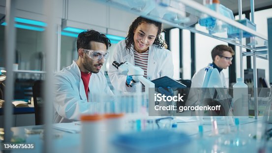 istock Modern Medical Research Laboratory: Portrait of Latin and Black Young Scientists Using Microscope, Digital Tablet, Doing Sample Analysis, Talking. Diverse Team of Specialists work in Advanced Lab 1346675635