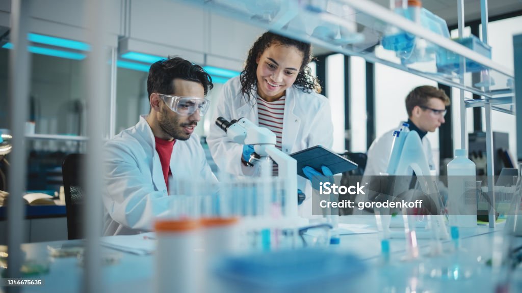 Modern Medical Research Laboratory: Portrait of Latin and Black Young Scientists Using Microscope, Digital Tablet, Doing Sample Analysis, Talking. Diverse Team of Specialists work in Advanced Lab Laboratory Stock Photo