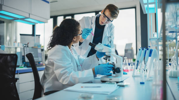 istock Modern Medical Research Laboratory: Portrait of Two Young Scientists Microscope, Doing Sample Analysis, Talking. Diverse Team of Multi-Ethnic Specialists work in Advanced Lab 1346675637