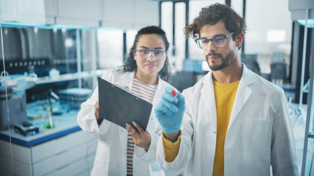 istock Medical Science Laboratory: Handsome Latin Male Scientist Writes Project Data Analysis on the Glass Board, His Black Female Colleague Checks Information on Clipboard. Young Scientists Solving Problems 1346675669