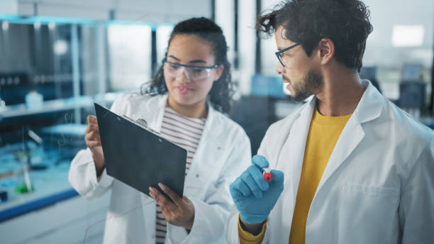 istock Medical Science Laboratory: Handsome Latin Male Scientist Doing Project Data Analysis, His Black Female Colleague Checks Information on Clipboard. Young Scientists Solving Problems 1346675671