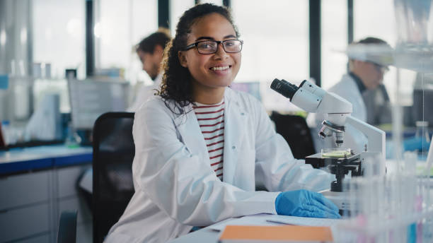 의학 연구소: 아름다운 흑인 과학자는 현미경을 사용 하 여, 카메라를 보고 매력적으로 미소. 젊은 생명 공학 과학 전문가, 기술적으로 진보 된 장비를 사용 하 여. - teamwork medical research science women 뉴스 사진 이미지