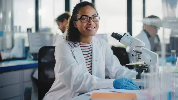 Photo of Medical Science Laboratory: Beautiful Black Scientist is Using Microscope, Looking at Camera and Smiling Charmingly. Young Biotechnology Science Specialist, Using Technologically Advanced Equipment.