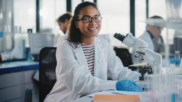 laboratório de ciência médica: belo cientista negro está usando microscópio, olhando para a câmera e sorrindo encantadoramente. jovem especialista em ciência da biotecnologia, utilizando equipamentos tecnologicamente avançados. - aluno de científico - fotografias e filmes do acervo